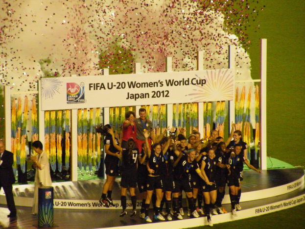 Frauenfußballspielerinnen in schwarzer Uniform feiern auf einer Bühne vor einem Publikum, mit einem Hintergrundschild und Dekorationsgegenständen auf Gras.