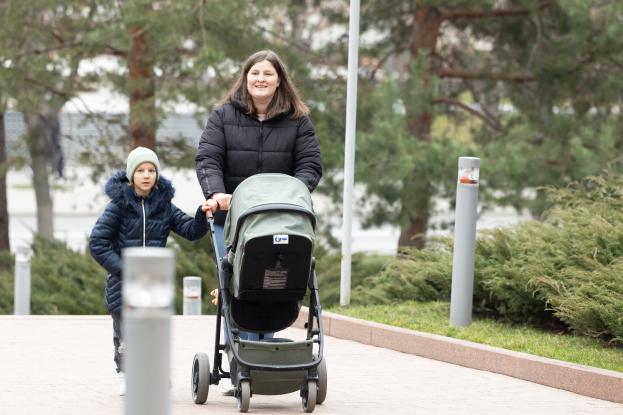 Eine Frau schiebt einen Kinderwagen mit einem Kind eine von Pfählen, Gras, Pflanzen und Bäumen gesäumte Straße entlang.
