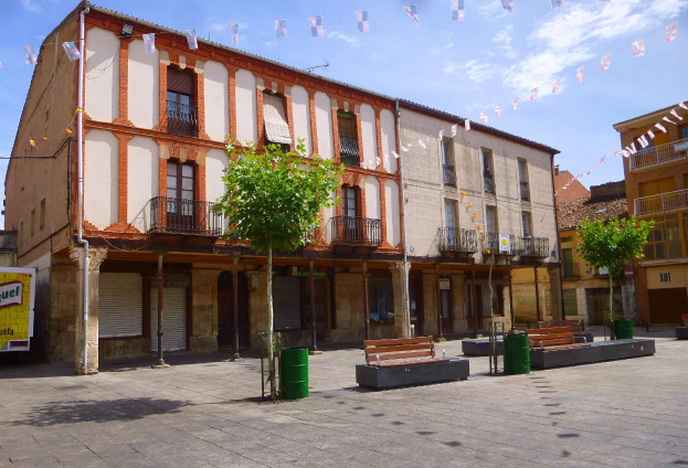 Ein Platz mit Gebäuden, Fenstern, Geländern, Bäumen, Bänken, Mülltonnen, Pfählen, Drähten und dekorativen Fahnen unter einem klaren blauen Himmel.