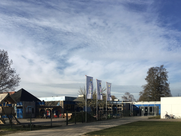 Großes Gebäude mit Metallzaun, Rasenfläche, Weg, Bäumen, Schildern, Fahrrad, benachbarten Häusern und bewölktem Himmel, identifiziert als neuer Spielplatz an der Schule.