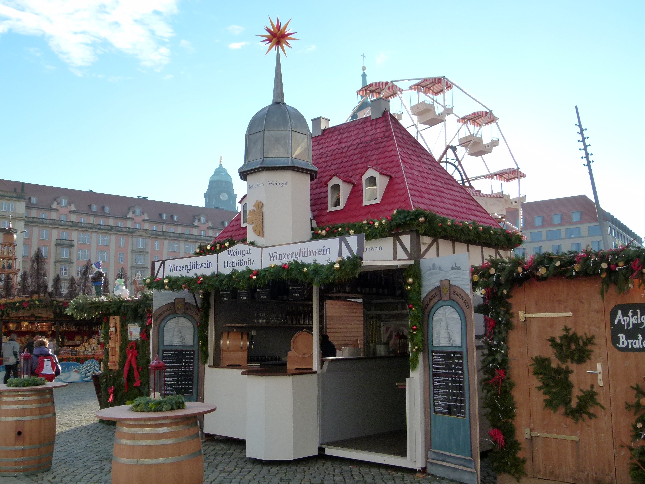 Ein kleines Gebäude mit rotem Dach und einem Riesenrad davor, umgeben von Festdekorationen und Menschen, mit Gebäuden, Bäumen und einem bewölkten Himmel im Hintergrund auf einem Oktoberfest-Event in München, Deutschland.