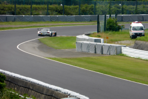 Ein Rennauto fährt eine Rennstrecke entlang, im Hintergrund ist ein Polizeiauto zu sehen, umgeben von Gras, Pflanzen, Bäumen und Absperrungen, mit einem Fahrzeug auf der rechten Seite und Text am unteren Bildrand.