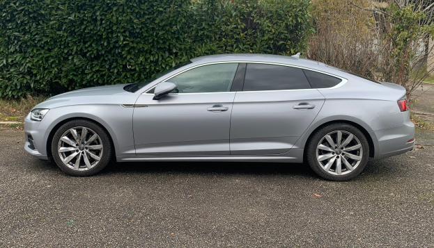 Grauer Audi A5 Sportback 2.0 TDI Quattro Kombi auf einer Straße mit Bäumen und einem Gebäude im Hintergrund.