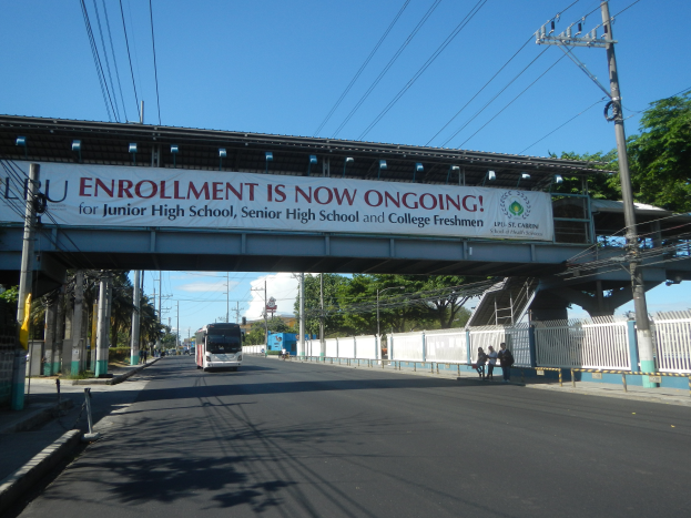 Ein Bus fährt unter einer Brücke hindurch, auf der ein Schild mit der Aufschrift "Jetzt Anmelden" steht, während Menschen auf dem Gehweg darunter gehen, mit Strommasten und Drähten an der Straße und Bäumen und einem klaren blauen Himmel im Hintergrund.