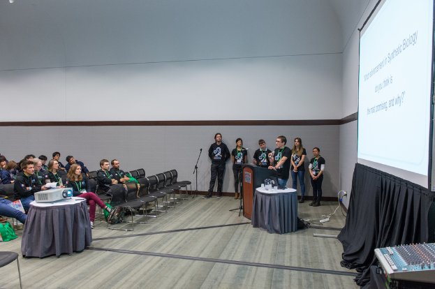 Eine Person spricht in ein Mikrofon auf einer Versammlung, mit Zuschauern und Teammitgliedern mit ID-Karten dahinter, einer Präsentation und einem Videoplayer in einem leeren Büroraum.