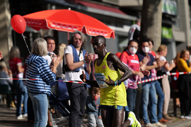Ein Mann in einem gelben T-Shirt und Laufschuhen sprintet durch einen Marathon, während ihn eine Menge im Hintergrund anfeuert, mit einem Schirm und einem Kinderwagen im Hintergrund.