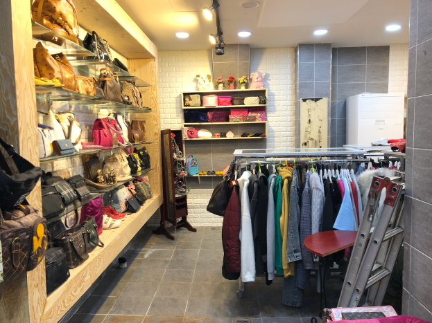 Ein Boutique-Innenraum mit Kleidung an Bügeln, Taschen und Handtaschen links, einem Tisch und einem Stuhl im Vordergrund und Deckenlampen.