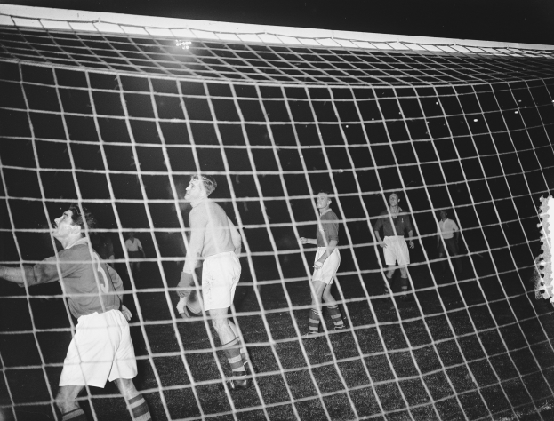 Eine Gruppe von Männern, die vor einem Tor mit Netz spielen, im Hintergrund beleuchtet von einem Licht, in Schwarz-Weiß.