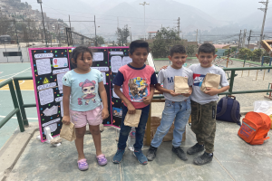 Gruppe von Kindern auf einem Basketballfeld, die Papierbeutel halten, mit Gegenständen rechts auf dem Boden und einer Tafel links, vor einem Hintergrund aus Zaun, Pfosten, Straßenlaternen, Bäumen, Gebäuden, Hügeln und Himmel.