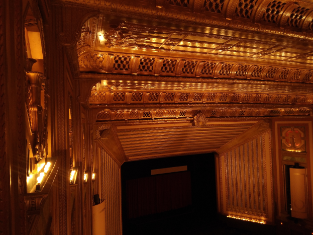 Ein prächtiger Theatersaal mit dunklem Hintergrund, beleuchtet von Lichtern auf beiden Seiten, mit einer kunstvoll gestalteten Decke.