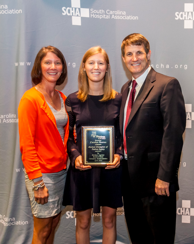 Drei Menschen, zwei Frauen und ein Mann, stehen zusammen. Der Mann trägt einen schwarzen Anzug, eine Frau trägt ein orangefarbenes und schwarzes Kleid, und die andere Frau trägt ein schwarzes Kleid und hält eine Trophäe in der Hand.