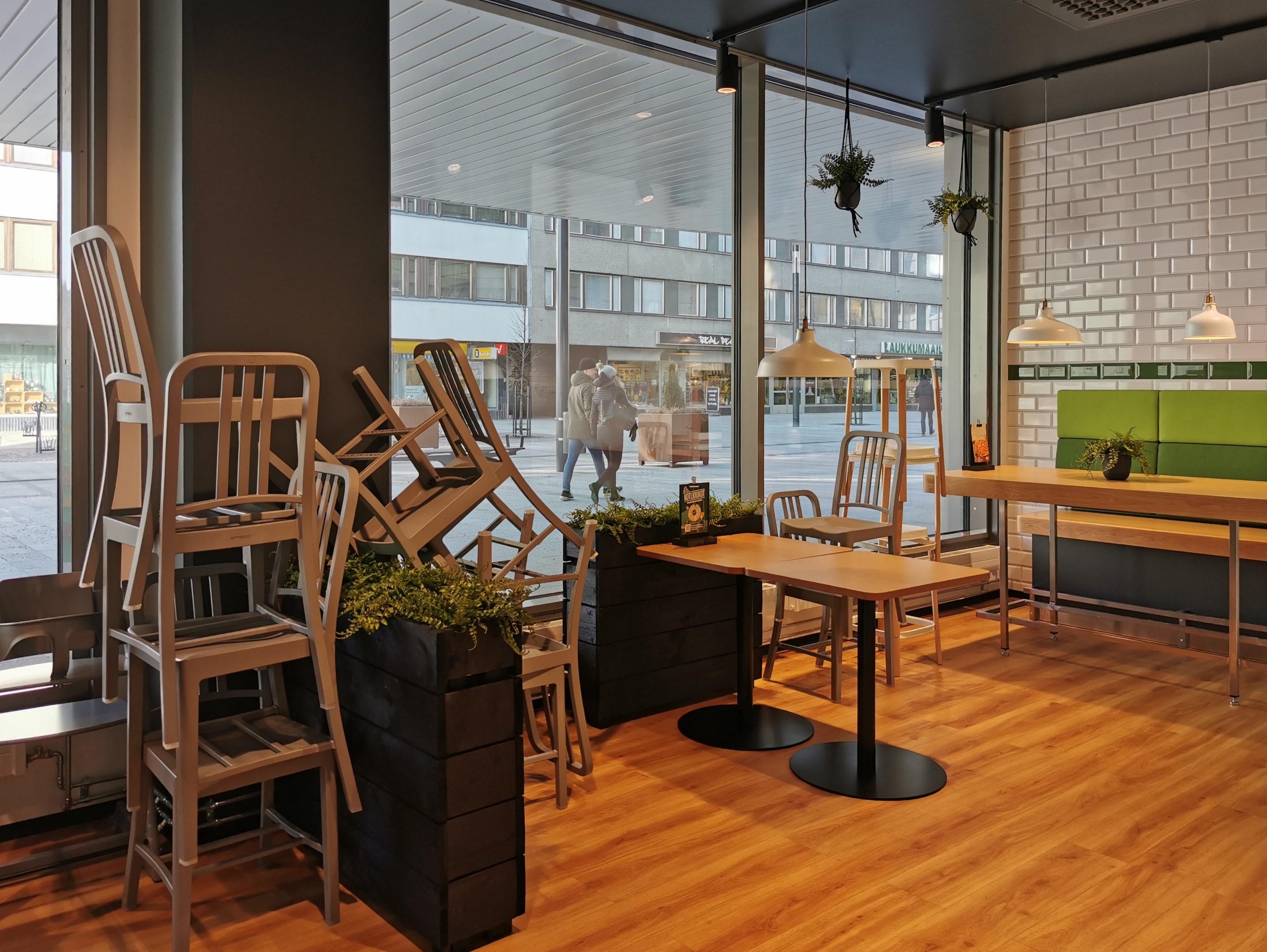Ein Restaurant-Innenraum mit hölzernen Tischen und Stühlen vor einem großen Fenster, das auf eine Berliner Straße mit Fußgängern, Gebäuden und urbanen Elementen blickt.