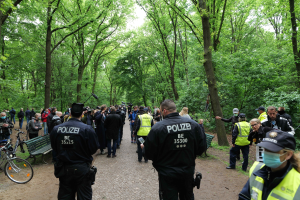 Polizeibeamte vor einer Menge bei einer Outdoor-Veranstaltung mit Fahrrädern und einer Bank im Vordergrund und Bäumen im Hintergrund.