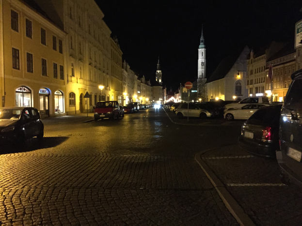 Eine nächtliche Straßenansicht mit parkenden Autos, Gebäuden mit Fenstern, Laternen, Schildern und einem Uhrenturm im Hintergrund unter einem dunklen Himmel.