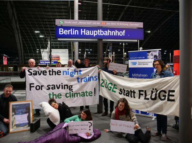 Eine Gruppe von Menschen sitzt auf dem Boden eines Bahnhofs und hält ein Banner mit der Aufschrift "Retten wir die Nachtzüge, um das Klima zu retten", umgeben von Kissen, einem Foto Rahmen und anderen Gegenständen, mit Plakaten, Säulen, Lampen und Geländern im Hintergrund.