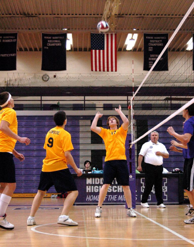 Menschen stehen auf einem Spielfeld mit Netz und Ball, tragen Kleidung und Schuhe, mit einem Landesflagge, Wand und Postern mit Text.