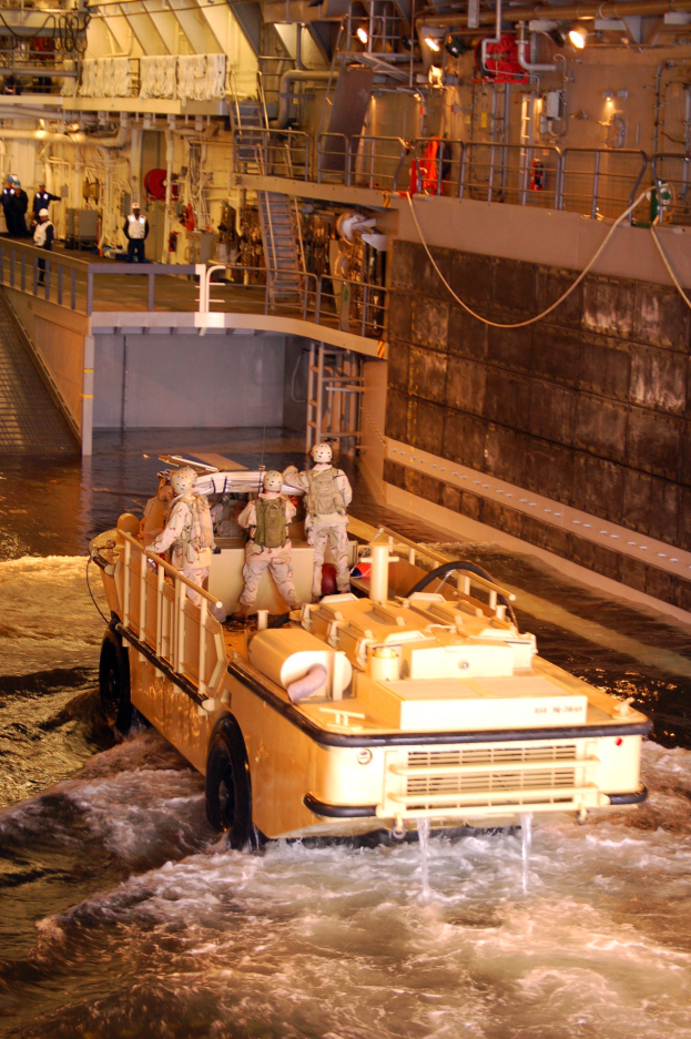 Amphibisches Fahrzeug mit Helmträgern durch Wasser in der Nähe einer Wand, mit Geländern, Lampen, Rohren und Menschen im Hintergrund.