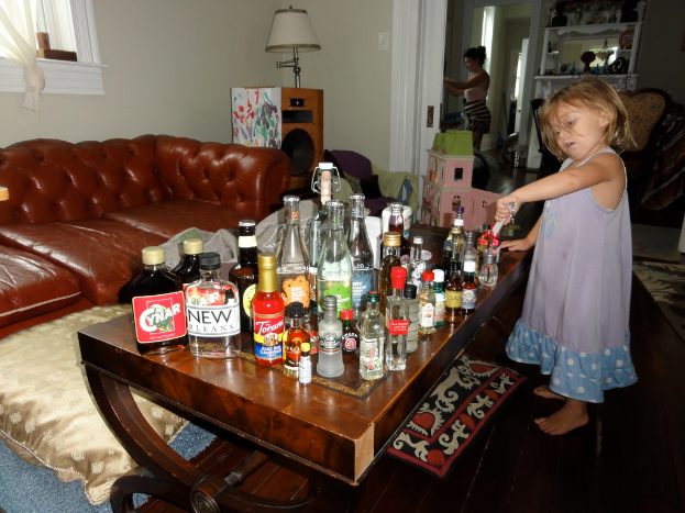 Ein kleines Mädchen steht in einem Wohnzimmer vor einem Tisch mit vielen Glasflaschen, einer Couch, einem Lautsprecher mit einer Lampe darauf und einer Frau im Hintergrund.