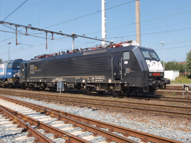 Ein schwarzer Zug, der auf Schienen neben einem Wald mit Strommasten und -leitungen, Bäumen, Gebäuden und einem klaren blauen Himmel fährt.