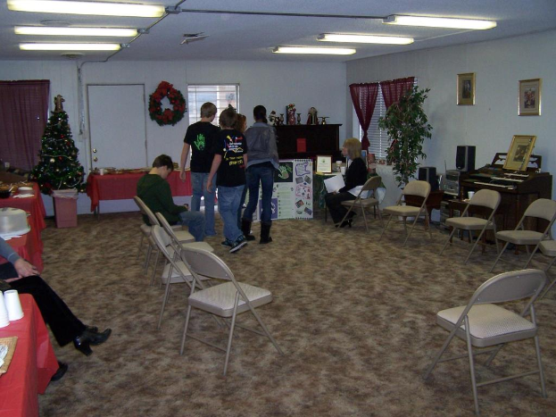 Ein Raum mit mehreren Stühlen und Tischen, einige Personen stehen, andere sitzen, Tassen und Gefäße auf den Tischen, eine Wand mit einem Strauß, Vorhang, Tür, Foto Rahmen und Fenstern, ein Weihnachtsbaum, ein Piano auf der rechten Seite und Röhrenleuchten an der Decke.