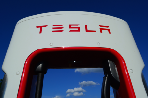 Eine weiße und rote Tesla-Ladestation mit dem Wort "Tesla" darauf, umgeben von Bäumen und einem blauen Himmel mit Wolken im Hintergrund.