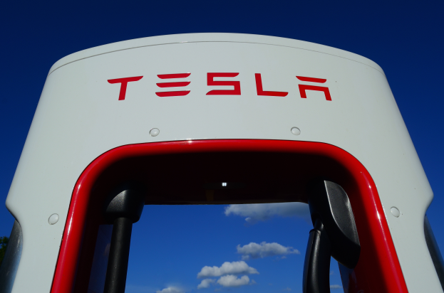 Eine weiße und rote Tesla-Ladestation mit dem Wort "Tesla" darauf, umgeben von Bäumen und einem blauen Himmel mit Wolken im Hintergrund.