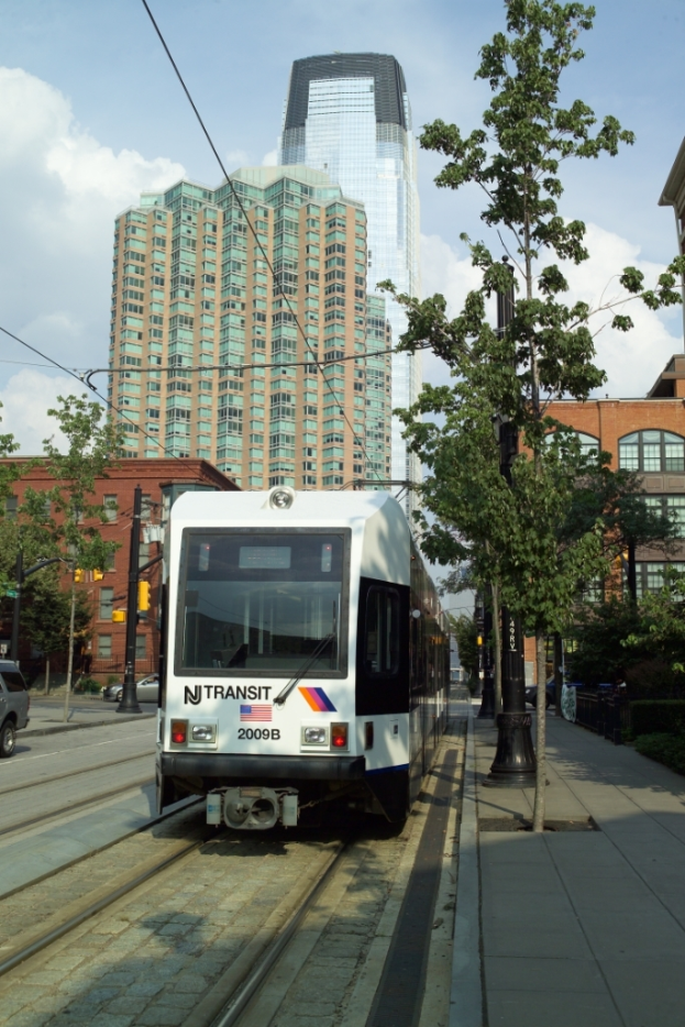 Ein Zug auf Schienen mit Oberleitungen, flankiert von B├Ąumen, Masten und Geb├Ąuden, mit einem Wolkenkratzer und Himmel im Hintergrund.