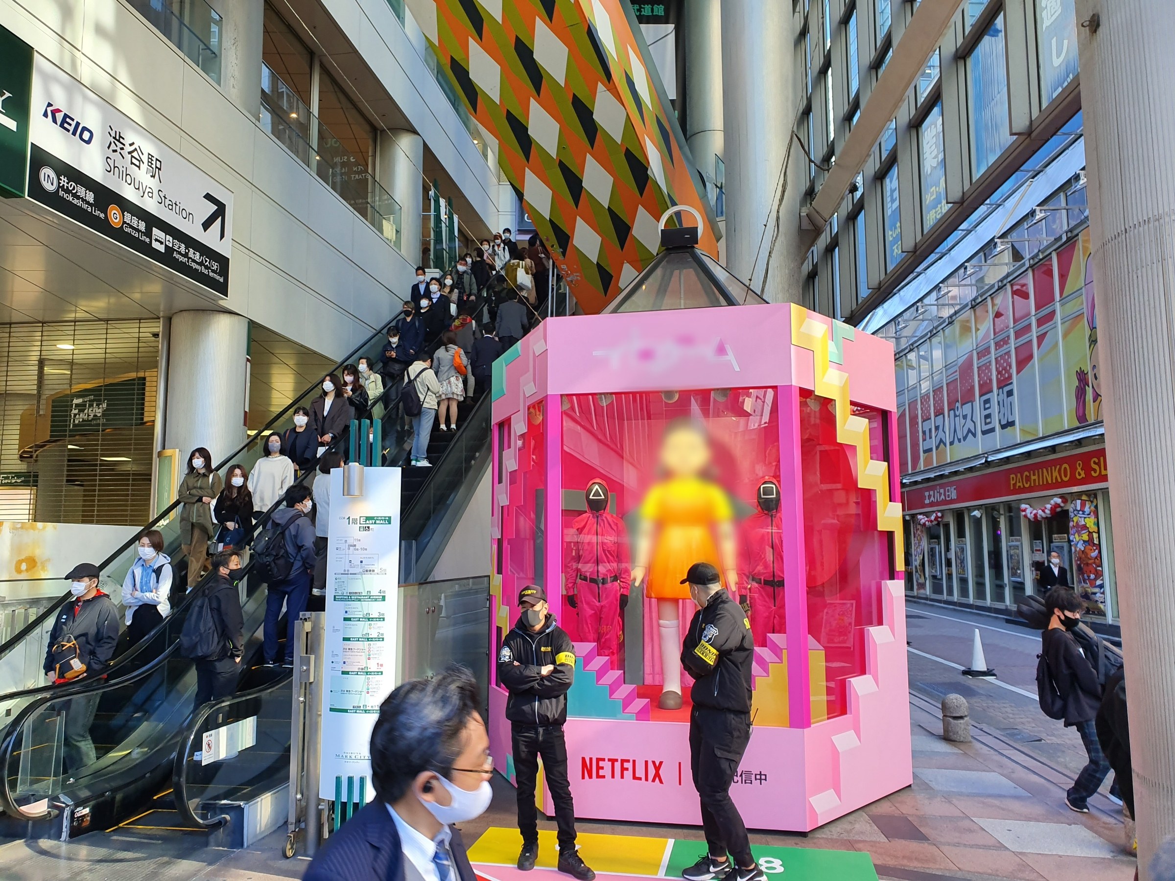 Eine Gruppe von Menschen steht vor einem Gebäude mit Glaswänden mit Texttafeln, einige tragen Masken, in der Nähe einer Rolltreppe; ein pinker Kasten mit Schaufensterpuppen zeigt "Netflix's new Netflix Booth in Tokyo."