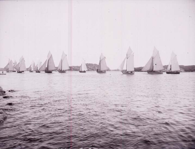 Schwarzes und weißes Foto von Segelbooten auf dem Wasser mit Hügeln im Hintergrund und einem klaren Himmel, beschriftet als Foto des New York Yacht Clubs.