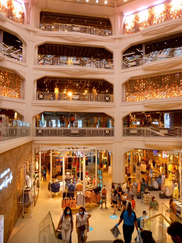 Ein belebter Einkaufszentrumsvorplatz mit Menschen, die Taschen tragen und Schaufensterpuppen mit Kleidern, umgeben von Geländern, Lampen und Wandtafeln.