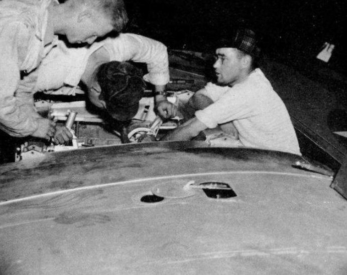 Drei Männer in Kappen arbeiten an einem schwarz-weißen Auto in einer Garage, umgeben von Werkzeugen und Reparaturmaterialien.