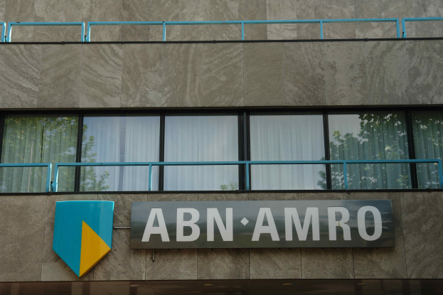 Ein Gebäude mit einer Tafel, auf der "ABN AMRO" steht, mit einer Tafel, die es als Hauptsitz angibt, einem Geländer und Glasfenstern mit Vorhängen, mit Text unten, der eine Strafe von 1,5 Milliarden Dollar durch die Europäische Union erwähnt.