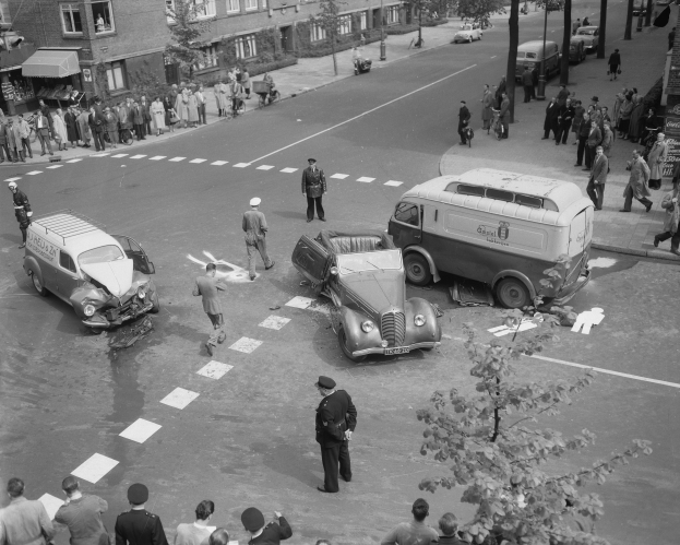 Eine Schwarz-Weiß-Szene eines Autounfalls an einer Straßenecke mit mehreren Fahrzeugen, darunter einem Lieferwagen, einer Gruppe von Menschen drumherum und Gebäuden, Bäumen, Masten und Texttafeln im Hintergrund.