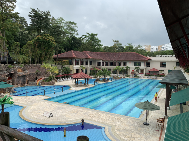 Großes Schwimmbad im Ritz-Carlton, Kota Kinabalu, mit Menschen, Sonnenschirmen, Stühlen, Tischen, Pflanzen, Bäumen, Felsen, Gebäuden und einem Schuppen auf der rechten Seite sowie einem sichtbaren Himmel im Hintergrund.