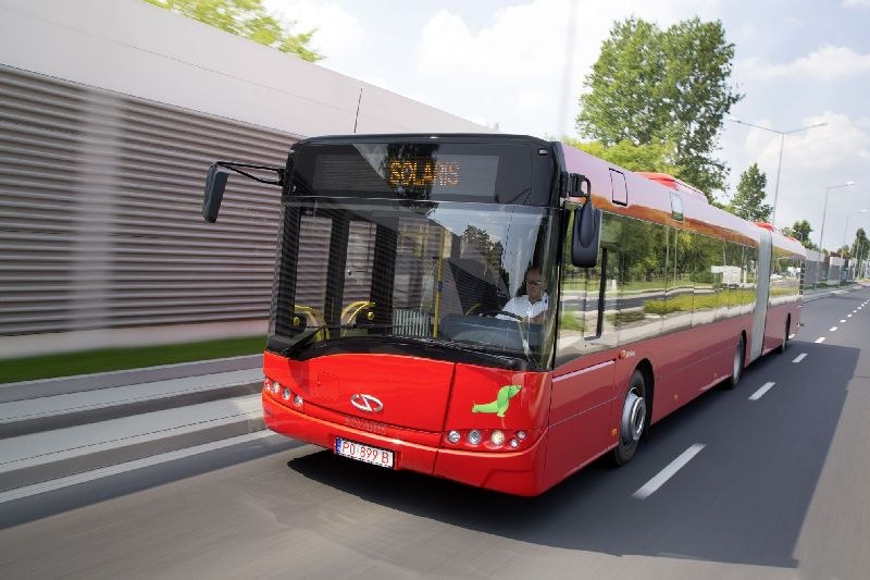 Roter Bus fährt auf einer Straße neben einem Gebäude, mit einer Person im Inneren, neben einer Wand, Bäumen, Laternen und einem bewölkten Himmel im Hintergrund.