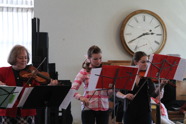 Drei Frauen, die auf der Bühne Geige spielen, während das Publikum zuhört, mit Notenpulten vor ihnen, einer Uhr an der Wand und einem weißen Vorhang im Hintergrund.