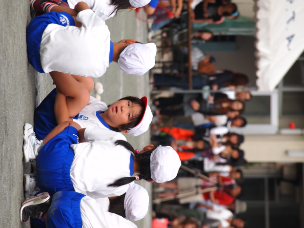 Kinder sitzen auf einer Straße im Vordergrund, Erwachsene stehen, ein Stativ, ein Zelt und ein Gebäude mit Glaswänden im Hintergrund.