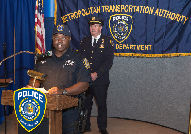 Zwei Polizeibeamte stehen vor einem Rednerpult, einer h\xlt Papiere und spricht in ein Mikrofon, mit einer Fahne, einem Tuch und einem Tisch in der N\xhe und einer Tafel mit Symbolen und Text im Hintergrund.