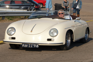 Ein Mann fährt einen weißen Porsche 356 Speedster die Straße entlang, umgeben von Menschen in bunten Kleidern, mit einem Geländer und einem schwarzen Müllimer zu beiden Seiten und weiteren Autos und einem Gebäude im Hintergrund.