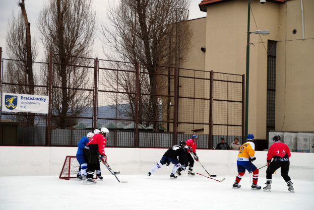 Personen beim Eishockey auf einem Eisstadion mit Gebäuden, Bäumen, einer Straßenlaterne, einem Namenskürzel und Zäunen im Hintergrund unter einem Himmel.
