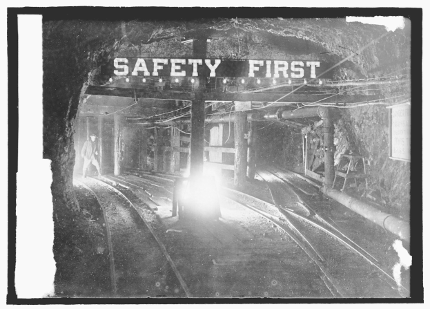 Schwaräweisses Foto eines Zuges, der durch einen Tunnel fährt, mit einer Person auf dem Gleis im Vordergrund, Masten und Lichter im Hintergrund und einem "Sicherheit geht vor"-Schild.