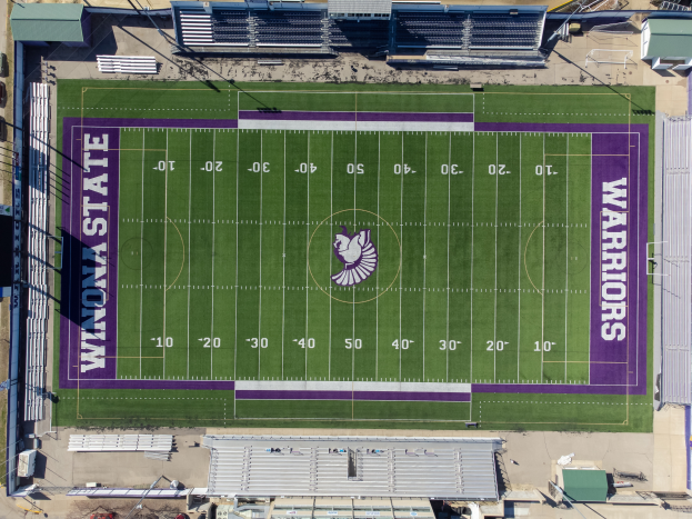 Eine Luftaufnahme eines Fußballfeldes mit violetten und weißen Markierungen, umgeben von Gebäuden, Fahrzeugen und Polen, die das Heimfeld der Minnesota State University Footballmannschaft zeigt.