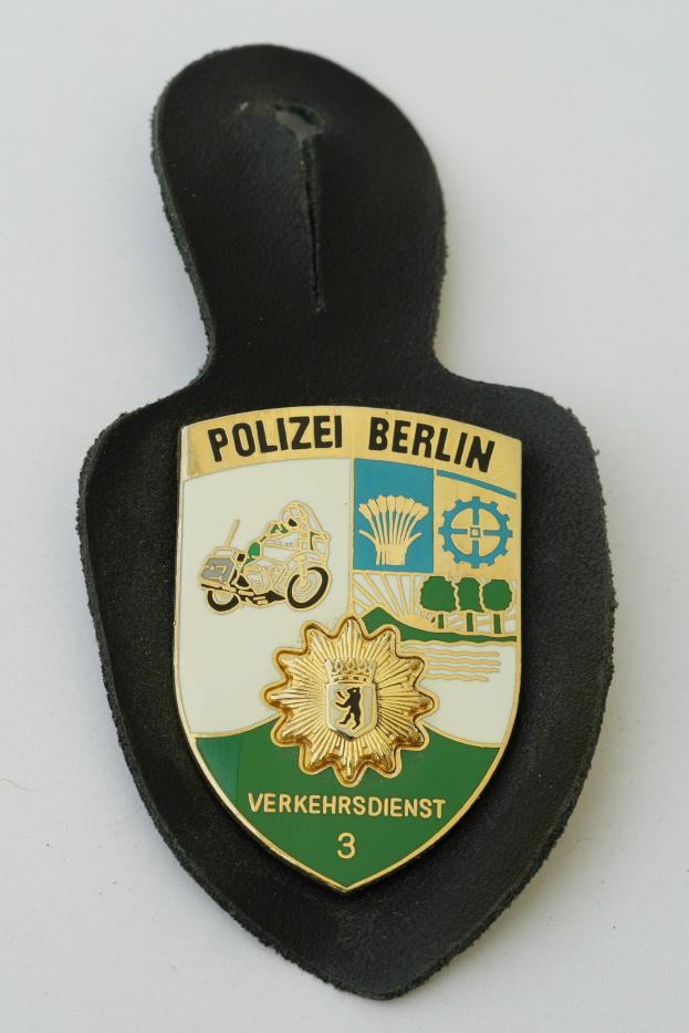 Ein schildförmiges Polizeisiegel mit der Aufschrift "Polizei Berlin" in schwarzer Fettschrift auf einem blauen Hintergrund mit einem weißen Stern und Rahmen, das auf einer Oberfläche liegt.