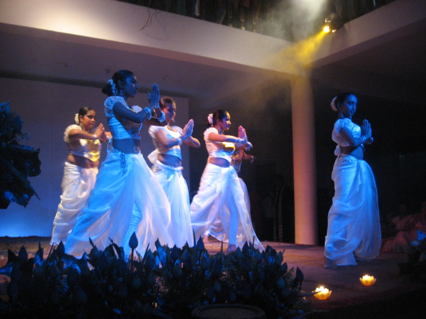 Eine Gruppe von Menschen, die auf einer Bühne mit Pflanzen davor tanzen und eine andere Gruppe von Menschen zuschauen.