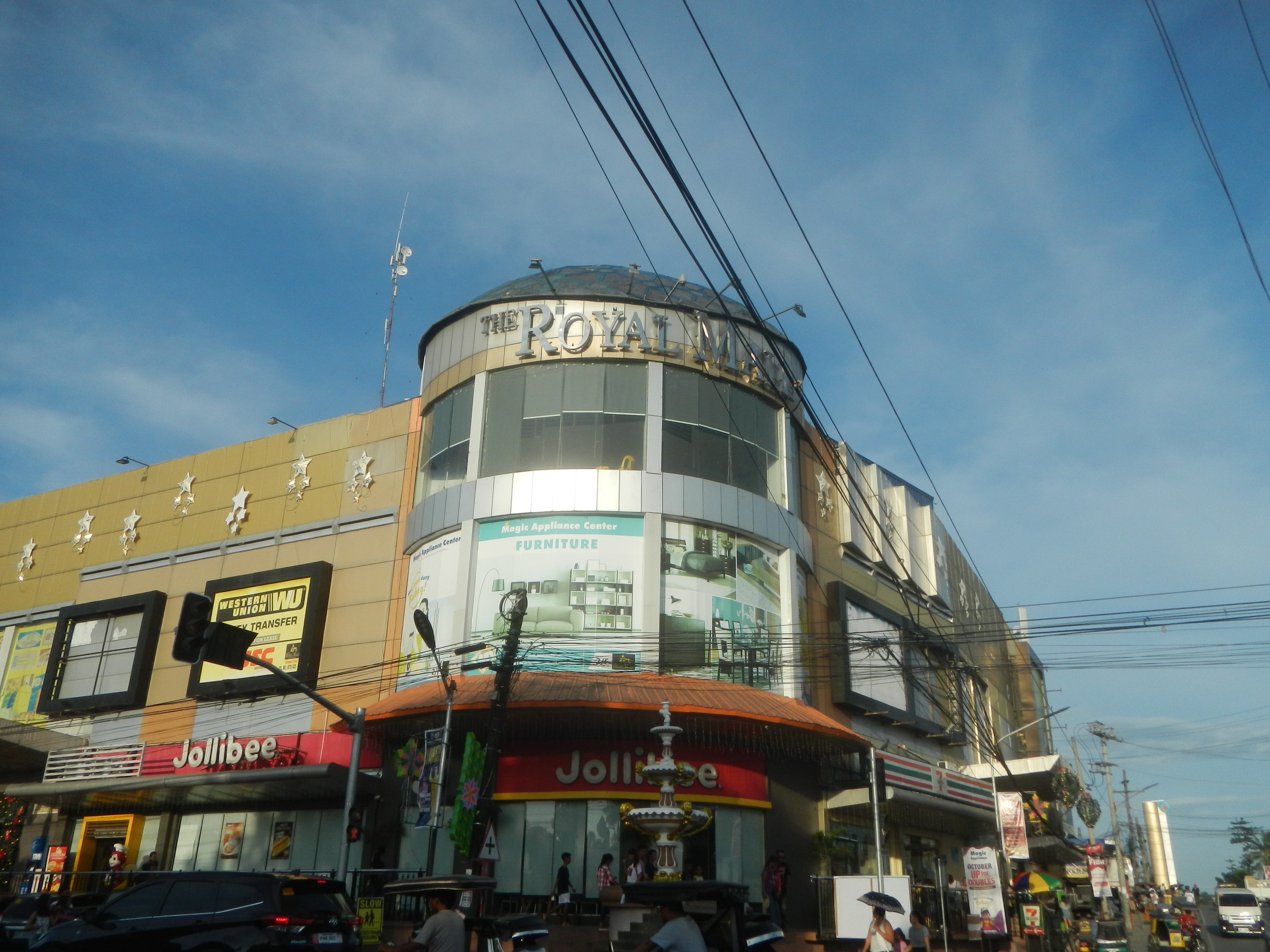 Eine belebte Stadtstraße mit fahrenden Fahrzeugen, Fußgängern auf dem Gehweg und dem Royal Mall-Einkaufszentrum im Hintergrund, umgeben von parkenden Autos, Bäumen, Strommasten und Drähten unter dem Himmel.
