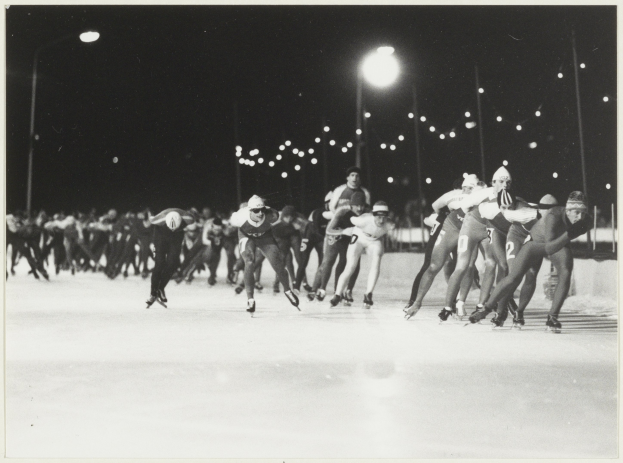 Eine Gruppe von Menschen, die auf einer Eisbahn im Schein von Straßenlaternen im Hintergrund nachts Schlittschuh laufen, auf einem Schwarz-Weiß-Foto.