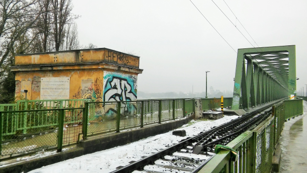 Eisenbahnschiene mit Graffiti an der Seite, umgeben von Gelöndern, Pfölen, Lichtern, Drähten und Bäumen, mit Schnee bedeckt und sichtbarem Himmel im Hintergrund.