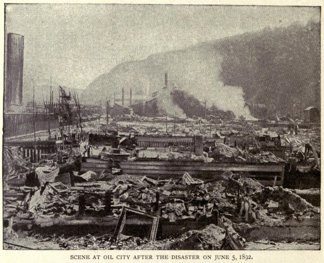 Schwarzes und weißes Foto einer Stadt nach einer Katastrophe am 5. Juni 1892, das Gebäude, Mäste, Rauch, einen Hügel im Hintergrund und Text unten zeigt.