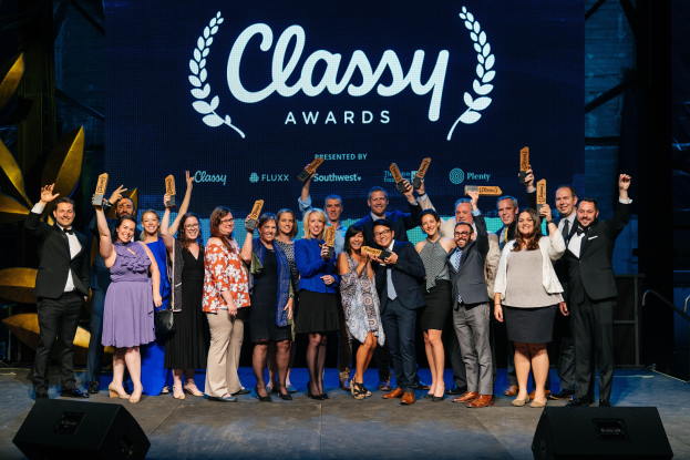 Eine Gruppe von Menschen auf einer Bühne, die lächeln und Auszeichnungen halten, mit Lautsprechern unten und einem Hintergrundbild mit "Classy Awards 2019".