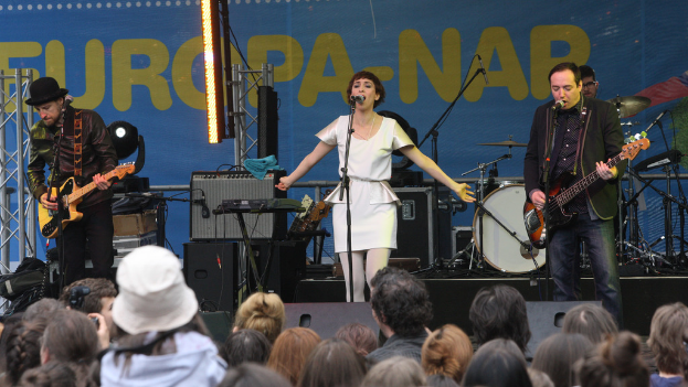 Eine Bühne mit vier Musikern: eine Frau in einem weißen Kleid singt in der Mitte, zwei Männer spielen Gitarren auf beiden Seiten, eine Menge vorne und Lautsprecher an der Wand im Hintergrund.